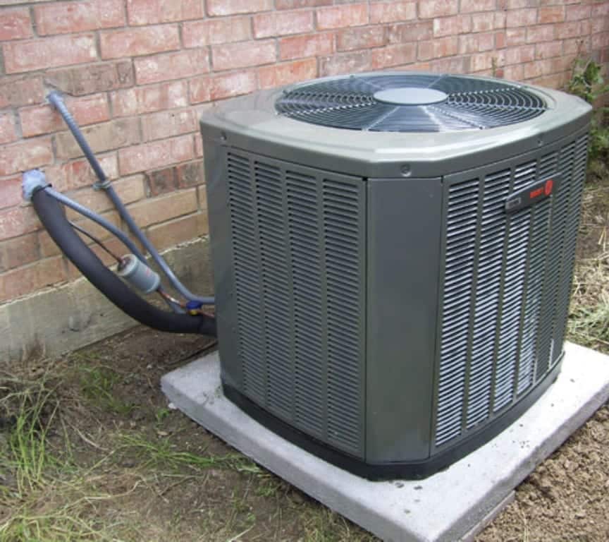 A central air conditioning unit is installed on a concrete pad outside a brick building, with connected pipes and wiring visible.