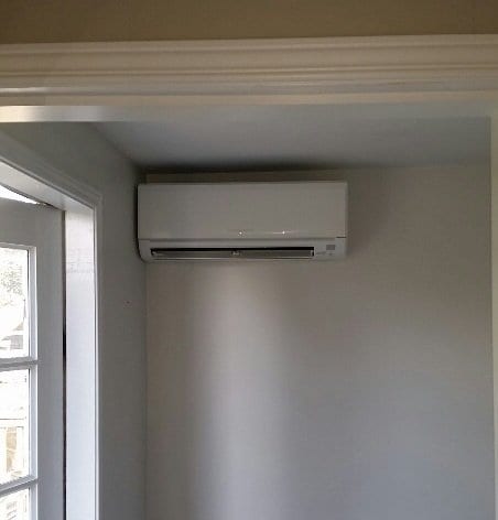 Wall-mounted air conditioning unit installed near the ceiling in a small, white room with part of a window visible on the left.