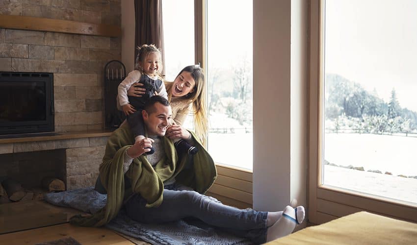 A family of three sits by large windows indoors, with snowy scenery outside. The child sits on an adult's shoulders, and all appear to be smiling and enjoying time together.