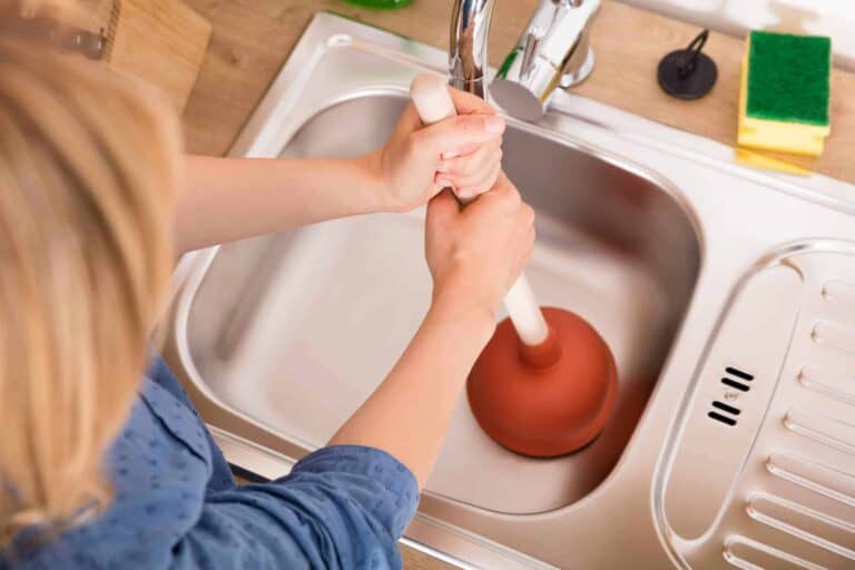 Person using a plunger in a kitchen sink, viewed from above, with cleaning supplies visible on the countertop.