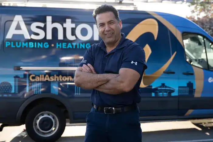Man in navy polo shirt standing with arms crossed in front of a blue Ashton Plumbing & Heating van in a parking lot.