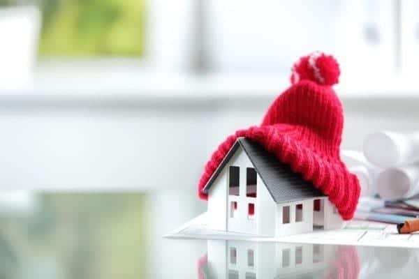 A small model house with a red knit winter hat placed on its roof, sitting on a glass surface with blurred papers in the background.