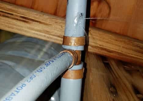 A close-up of a gray plastic water pipe with a small leak spraying water, held by brown brackets, near a wooden structure.