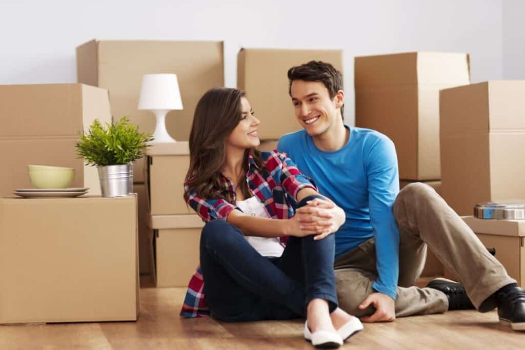 A young couple sits on the floor, smiling at each other, surrounded by cardboard moving boxes and household items in a room.