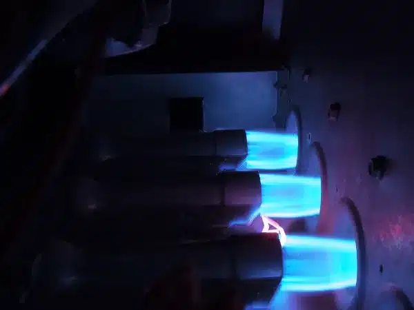 Close-up view of three blue gas flames burning inside a furnace, with metal burner tubes visible in a dark environment.
