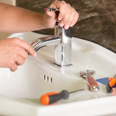Hands performing plumbing repair by installing a modern chrome faucet on a white bathroom sink, with various tools such as a wrench and screwdriver placed nearby.