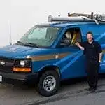 A man in dark clothing stands beside a blue utility van with a ladder mounted on the roof, parked on a paved surface.