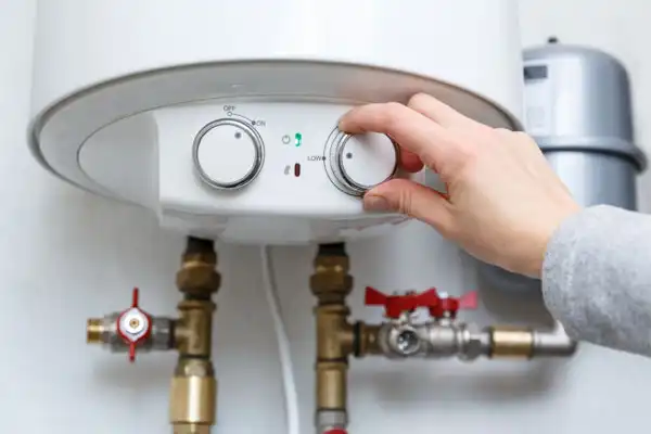 A hand adjusts the temperature dial on a tankless water heater mounted on a wall with visible pipes and valves below.