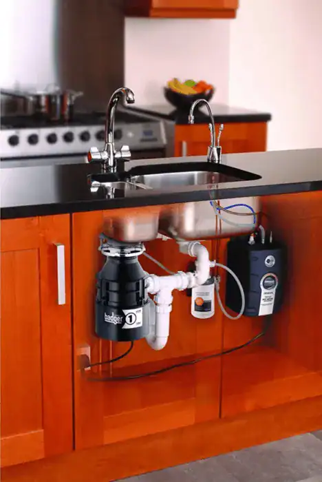 A kitchen sink with exposed plumbing showcases a garbage disposal unit ready for garburator repair, along with pipes, a water filter, and electrical components installed in a wooden cabinet.