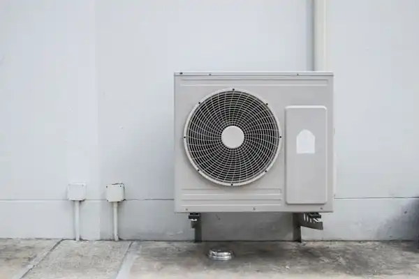 An outdoor air conditioning unit mounted on a wall above a concrete surface, with pipes running along the wall.