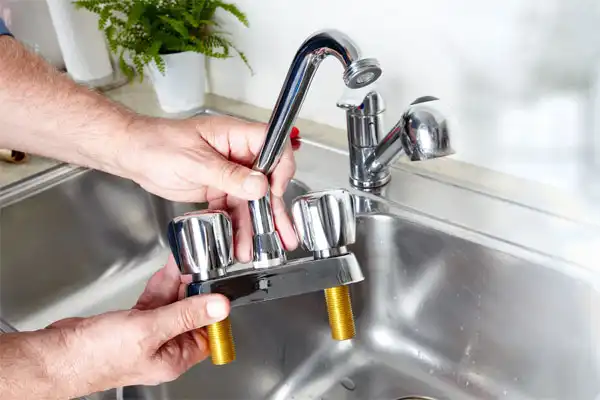 A person holding a kitchen sink faucet fixture above a stainless steel sink, preparing for installation or replacement.
