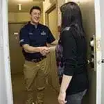 A man wearing a uniform shakes hands with a woman at the doorway of a building hallway.